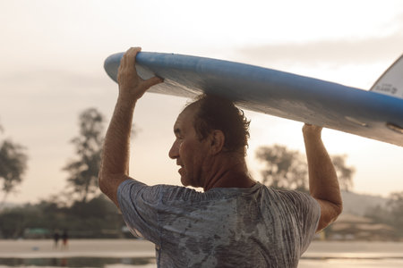 Rear view of mature surfer carrying surfboard on head and looking aside while coming out of sea to...