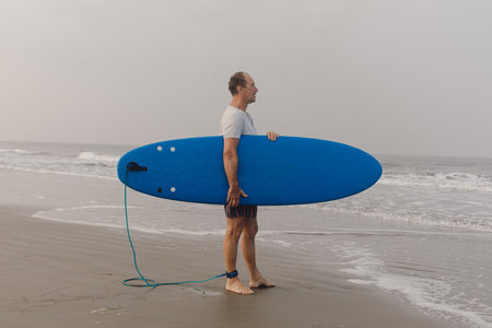 Mature athlete keeping blue surfboard under armpit standing on wet sandy shore in front of sea and looking at seascape.の写真素材
