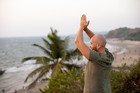 Calm vacationist folding palms together and lifting arms over head standing on hill near plants and looking at shore.の写真素材