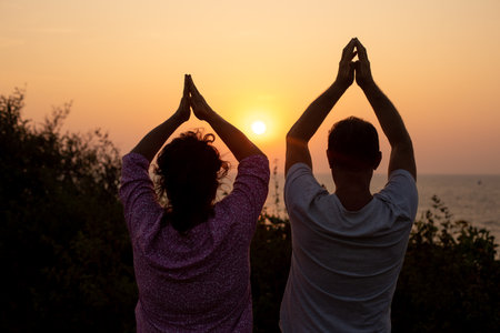 Rear view of woman and man folding palms together and lifting arms up watching at sunset, practicing yoga together.の写真素材