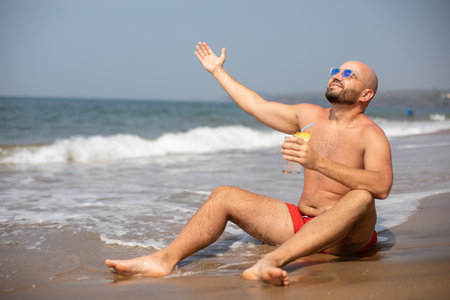 Topless vacationer in sunglasses with drink in hand lifting arm up and enjoying sun with closed eyes on wet seashore.の写真素材