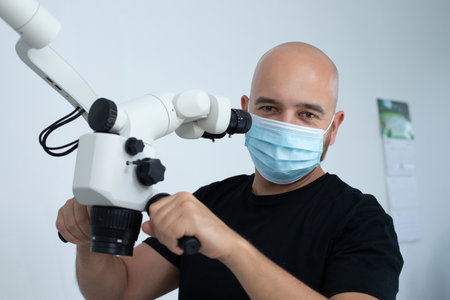 Bald male doctor in blue medical mask standing near dental microscope in clinic and holding hands on...