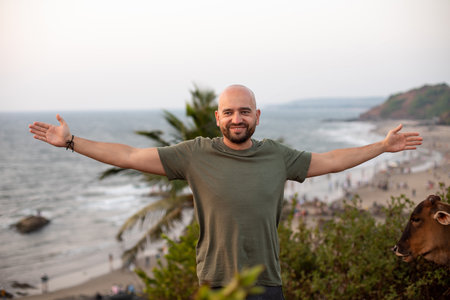 Happy man standing with arms wide open on green hill in front of seashore against seascape and enjoying vacation.の写真素材