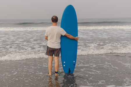 Rear view of mature man standing in waving water on seaside, holding blue surfboard and looking...