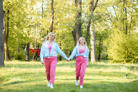 Charming blonde mom and her adult daughter, dressed in same clothes, walk hand in hand, step by step on walk in park.の写真素材