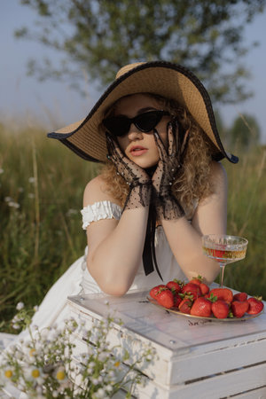 Strawberries, champagne. Gorgeous blonde in elegant hat and transparent black gloves on summer day.の写真素材
