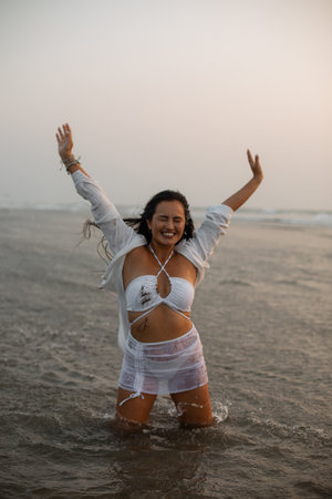 Portrait of young smiling Asian woman wearing white swimsuit, standing in water near beach at sunset, stretching hands.の写真素材