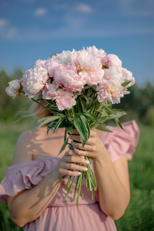 Unrecognizable young woman with flawless skin in sundress stands in nature with bouquet of peonies.の写真素材