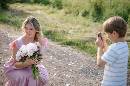 Preschooler son photographs his happy mother with bouquet of peonies on summer day in countryside.の写真素材