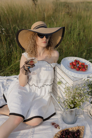 Picnic in nature on summer. Blonde in white dress, elegant hat drinks champagne with strawberries.の写真素材