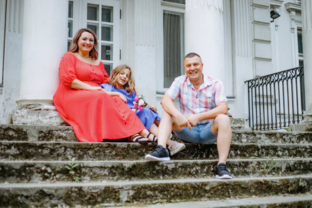 Beautiful woman is sitting on steps of wide staircase next to her charming daughter and husband.の写真素材