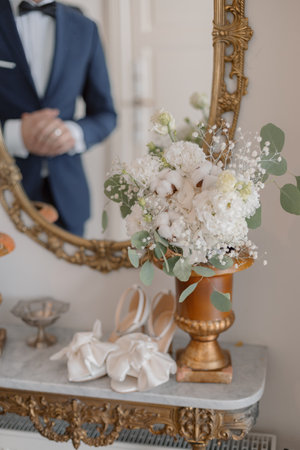 Elegant wedding shoes and snow-white bridal bouquet in vintage vase stand on old wall table.の写真素材