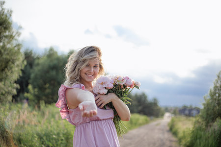 Lovely blonde with bouquet of pink peonies smiles coquettishly, holding out her hand calls for her.の写真素材