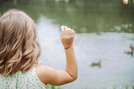 Unrecognizable girl in light dress stands with her back and feeds ducks near pond in nature.の写真素材