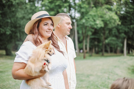 Beautiful woman in summer dress, hat with dog in her arms walks with her husband on summer in park.の写真素材