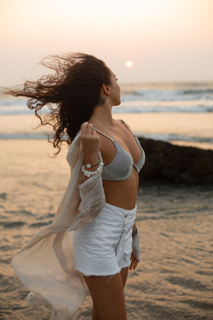 Side view of young woman wearing grey bra, white shorts, light shawl, standing in water on beach, looking at sunset.の写真素材
