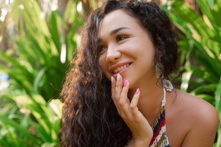 Portrait of young happy Asian woman with long curly hair sitting surrounded by green plants palm trees, touching lips.の写真素材