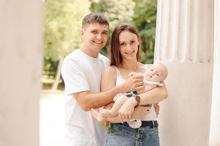 Young family on sunny summer day in nature, mom holds tiny baby in her arms, dad hugs them. Loans.の写真素材