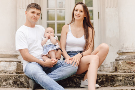 Beautiful married couple with little daughter in dads arms is sitting on summer day outside.の写真素材