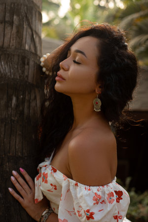 Portrait of young Asian woman wearing white blouse with red flowers, leaning with closed eyes on trunk of palm tree.の写真素材