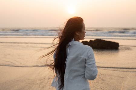 Back view of young Asian woman stand in water near beach, looking at waves reflecting sunset, enjoying holiday vacation.の写真素材