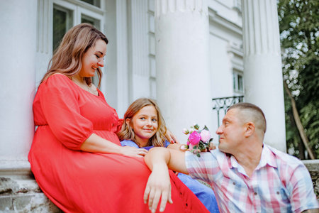 Charming woman in long bright red dress is sitting on steps next to her happy daughter and husband.の写真素材