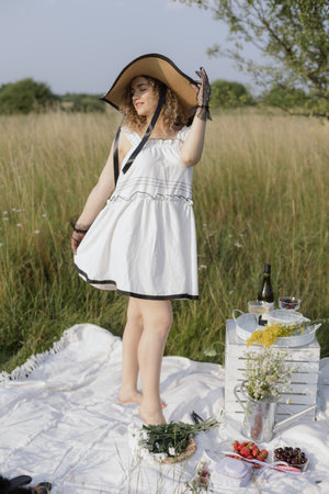 Elegant young girl in white dress, hat poses on summer day in nature, stands barefoot on blanket .の写真素材