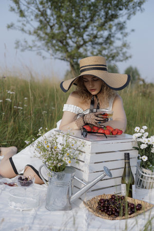 Concept of romantic picnic. Gorgeous blonde in elegant hat is enjoying champagne with strawberries.の写真素材