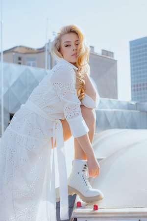 Flirtatious blonde in white dress poses, showing off her leg in stylish shoes, on roof of building. Modern women.の写真素材