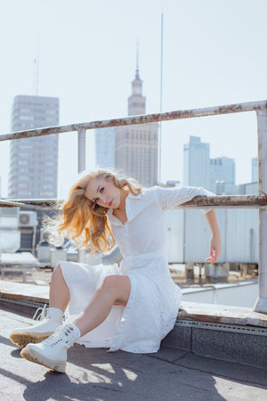 Urban fashion. Charming blonde in white dress poses, showing off comfortable stylish white shoes, on roof of building.の写真素材