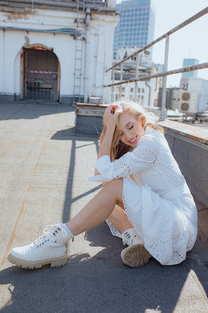 Positive emotions. Feminine blonde with long loose hair smiling, sitting on roof of city building on summer morning.の写真素材