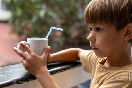 6-7-year-old boy is holding white mug with drink and drinking tube on summer day. Care of children.