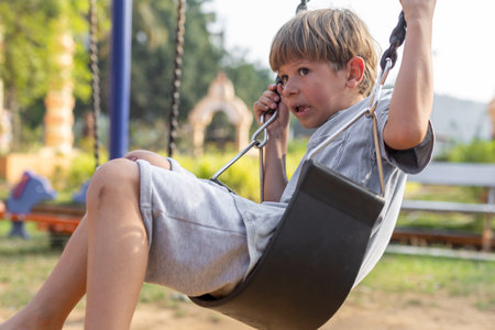 6-7-year-old boy in gray cotton T-shirt is sitting on swing in childrens playground and is afraid.