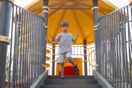 Cute boy in gray cotton T-shirt and shorts walks down stairs fromchildrens slide .の写真素材