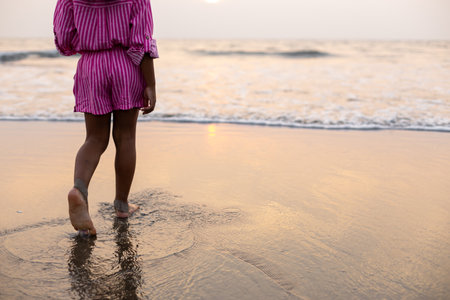 Layout for postcard. Unrecognizable girl walks barefoot through water to waves at sunset on beach.の写真素材