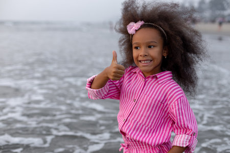 Lovely girl with loose curly hair enjoys walk on beach at sunset, smiles and shows her hand OK.の写真素材