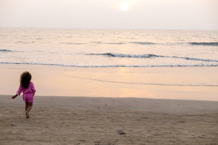 Unrecognizable girl with loose hair and dark skin runs along sandy beach to sea at sunset. Layout.の写真素材