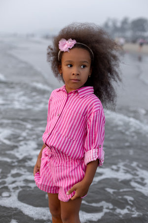 Vertical beautiful portrait of little girl with curly hair, standing in coming waves on beach.の写真素材