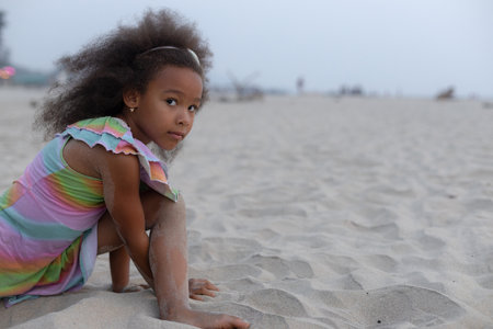 Charming little girl with dark skin is sitting on beach, looking at camera in summer morning.の写真素材