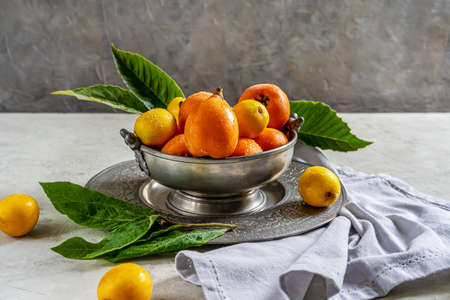 Yellow and orange Medlar fruits on antique tin bowl and plate with leaves over concrete background,...