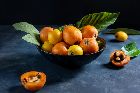 Yellow and orange Medlar fruits on plate with leaves over dark concrete background, two half of fruit on the tableの写真素材