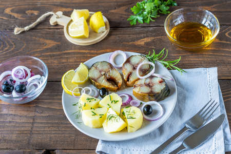 Cut smoked mackerel on plate with boiled potatoes, rings of onion, lemon, olives, oil, greens on...