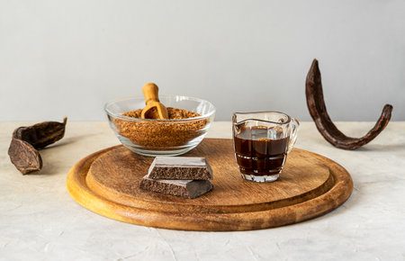 Carob molasses, flour, chocolate and carob pods on wooden cutting board with light grey background and copy spaceの写真素材