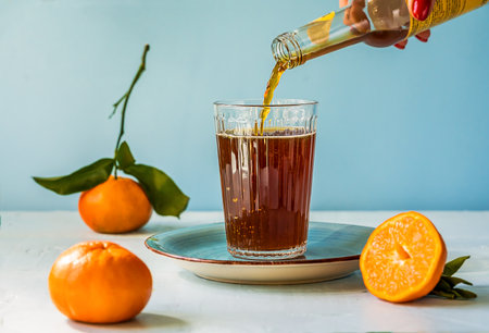 Chinotto refreshing citrus drink in a glass, womans hand holding bottle and pouring drink to the glass, light surface with blue backgroundの写真素材