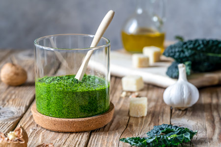 Homemade kale pesto served in a glass jar on wooden surface with grey background and basic ingredients - kale, walnuts, parmesan, garlic and olive oilの写真素材