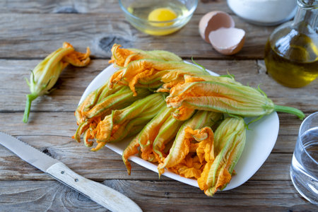 Zucchini flowers on a plate with basic ingredients to be fried in batter or making crepes. Wooden table, eggs, flour, olive oilの写真素材