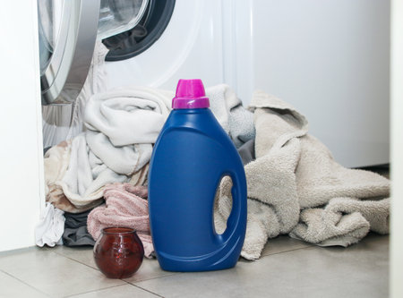 A blue bottle of liquid washing gel on grey floor near a washing machine with dirty clothの写真素材