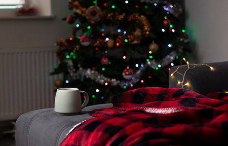 Close up of a mug with hot drink on a sofa with a red blanket, blurry background with a Christmas tree lightsの写真素材