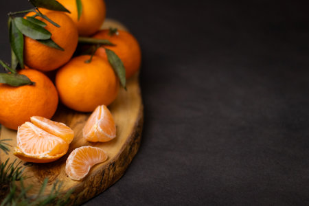 Close up of peeled mandarine slices and a pile whole mandarines on wooden plate, blurry green pine...