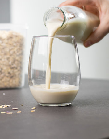 Close up of a female hand pouring oat milk into a glass on grey kitchen table. Vegan and non-dairy alternative milkの写真素材